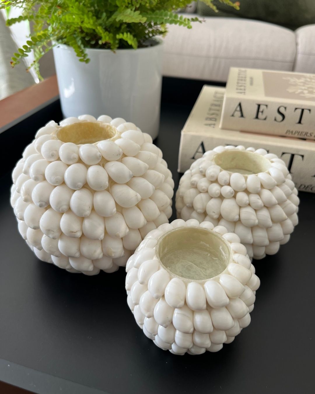 Three white textured candle holders on a dark surface with books and a plant in the background.