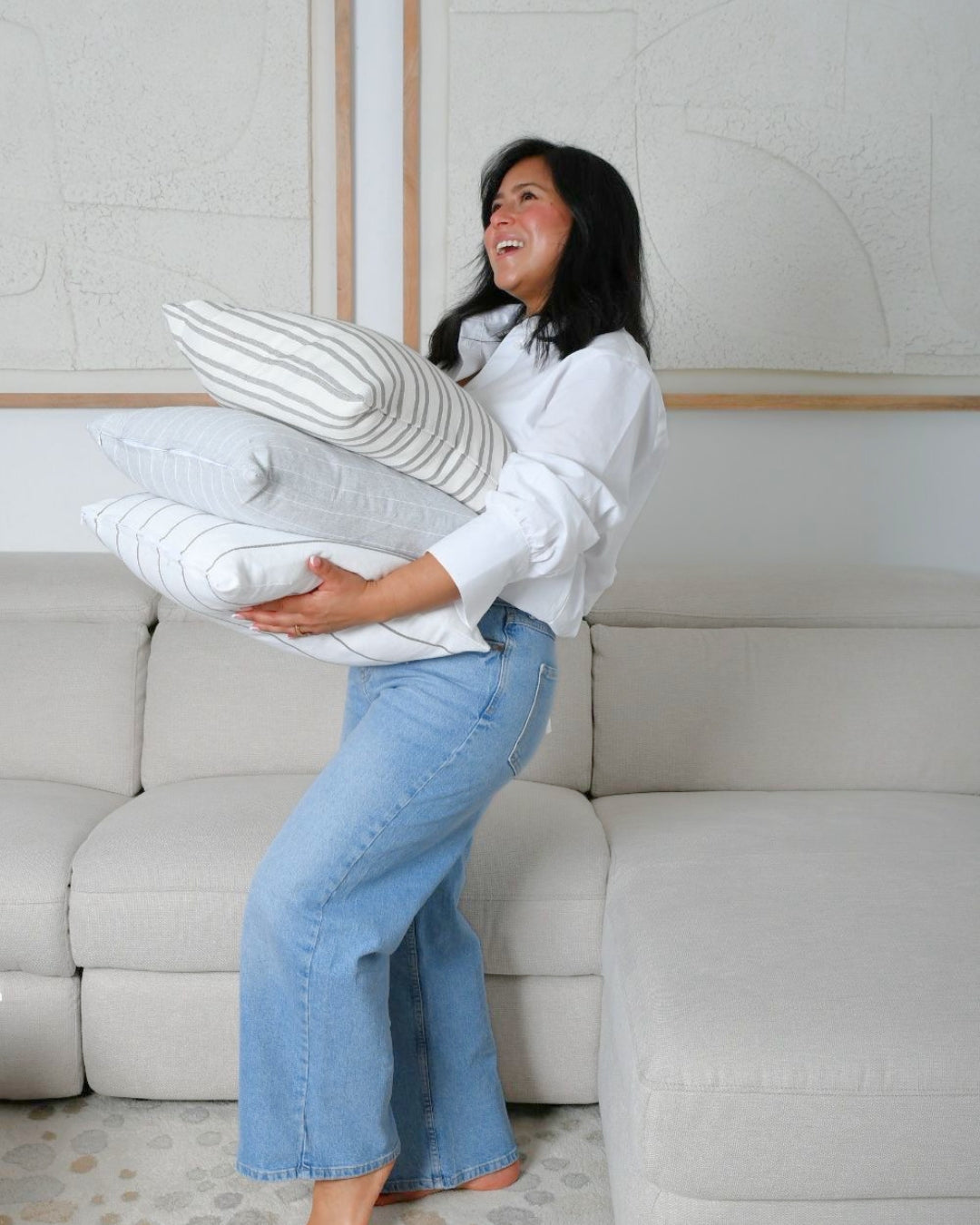 A woman in a white blouse and light blue jeans stands barefoot in a living room, holding three striped and solid throw pillows against a light beige sectional sofa.