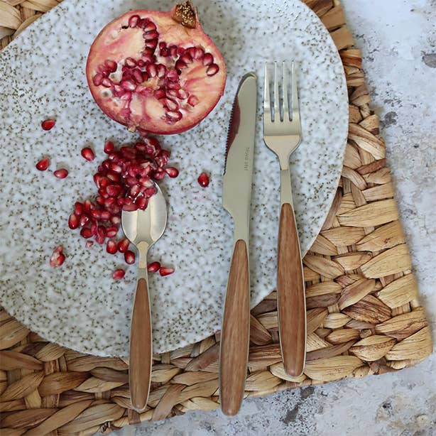 Cut pomegranate on a speckled plate with wooden cutlery on a woven mat. Suva wood-effect cutlery styled on a minimalist wooden table with neutral linens.