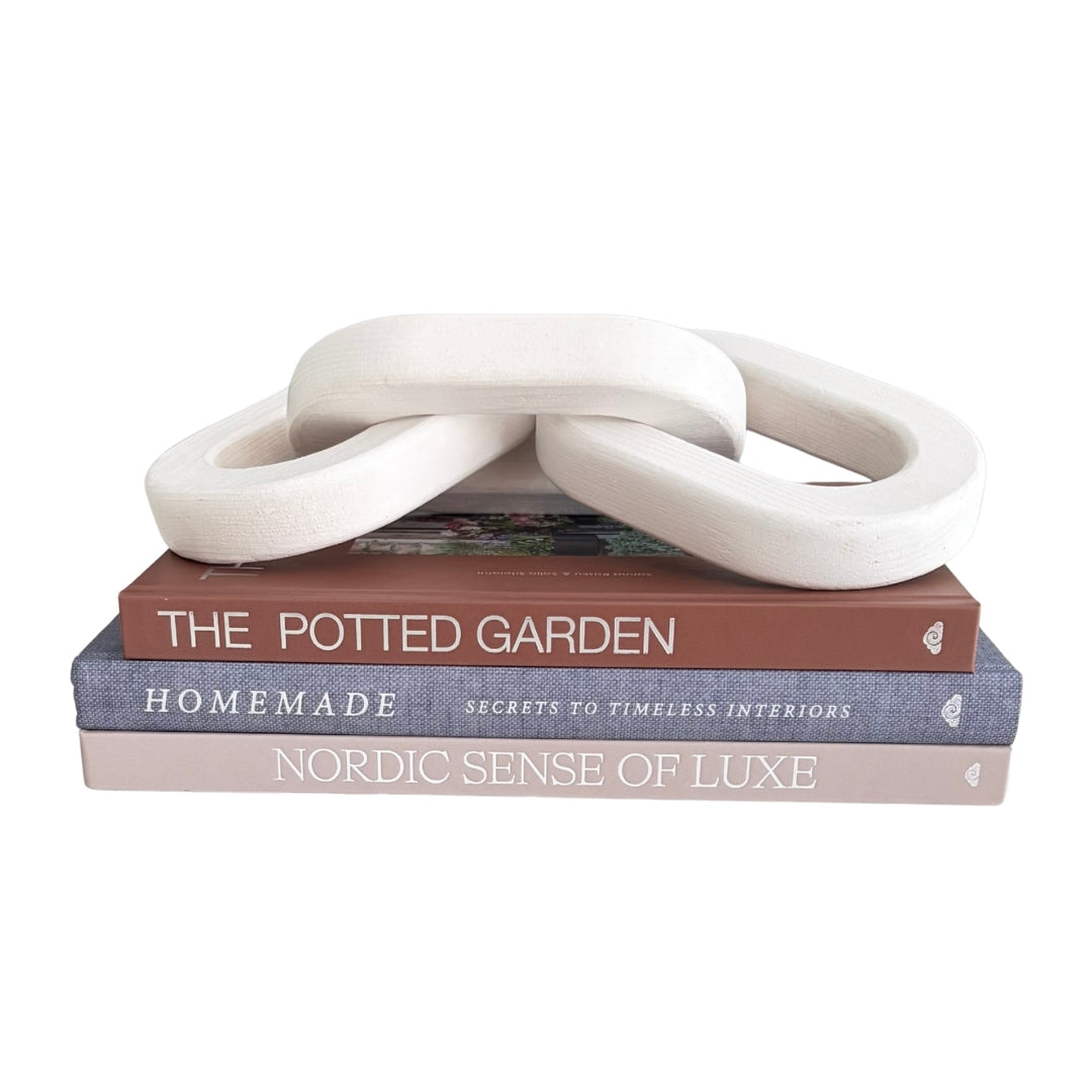 Three interlocking looped decorative objects in an off-white matte finish, displayed on top of a stack of three books titled 'The Potted Garden,' 'Homemade,' and 'Nordic Sense of Luxe' against a plain white background.