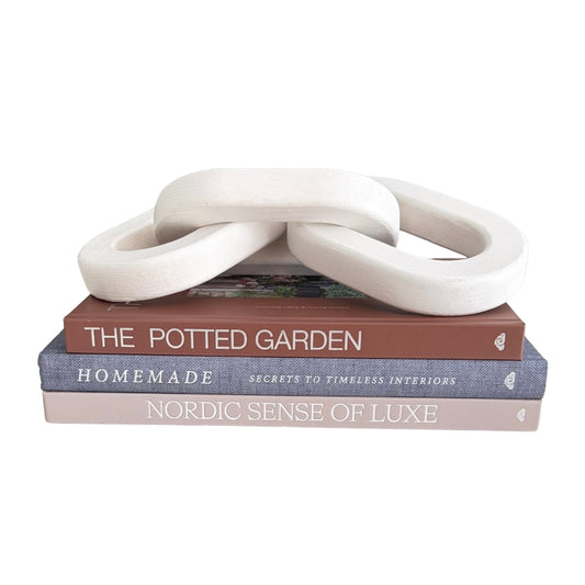 Three interlocking looped decorative objects in an off-white matte finish, displayed on top of a stack of three books titled 'The Potted Garden,' 'Homemade,' and 'Nordic Sense of Luxe' against a plain white background.