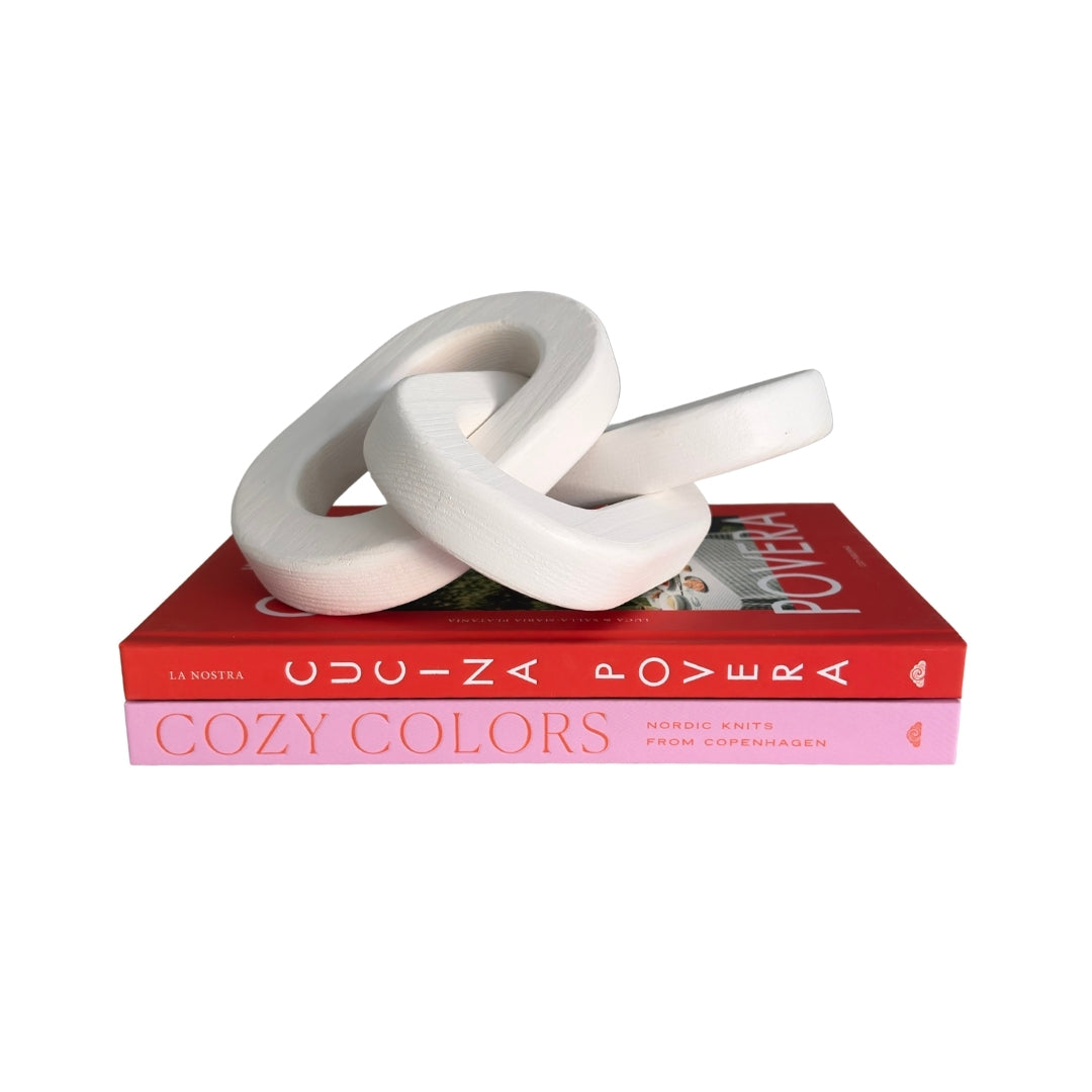 A white sculptural knot-like decorative object displayed on top of two stacked books, one red and one pink, against a plain white background.