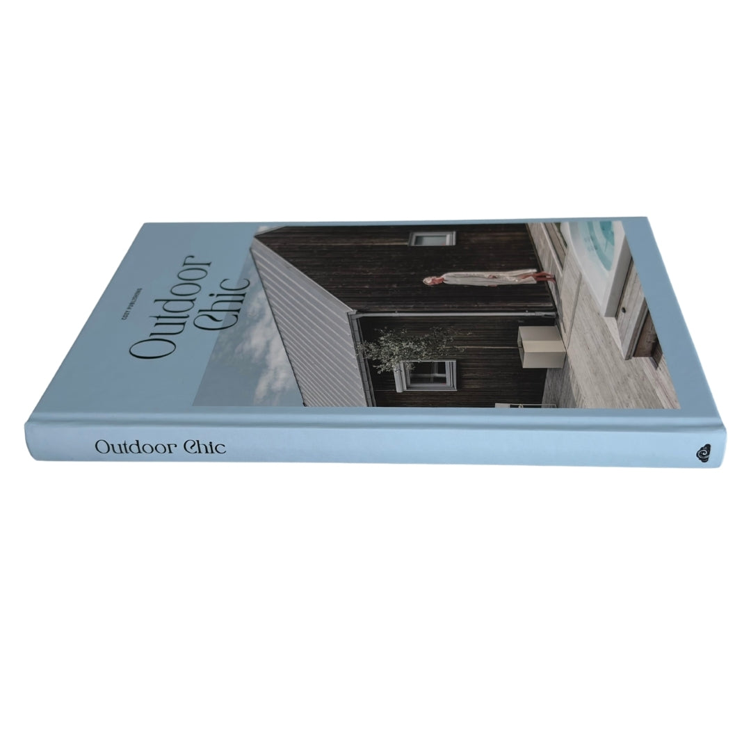 A hardcover book titled 'Outdoor Chic' with a light blue cover and spine, featuring a photograph of a rustic dark wooden house on the front cover.