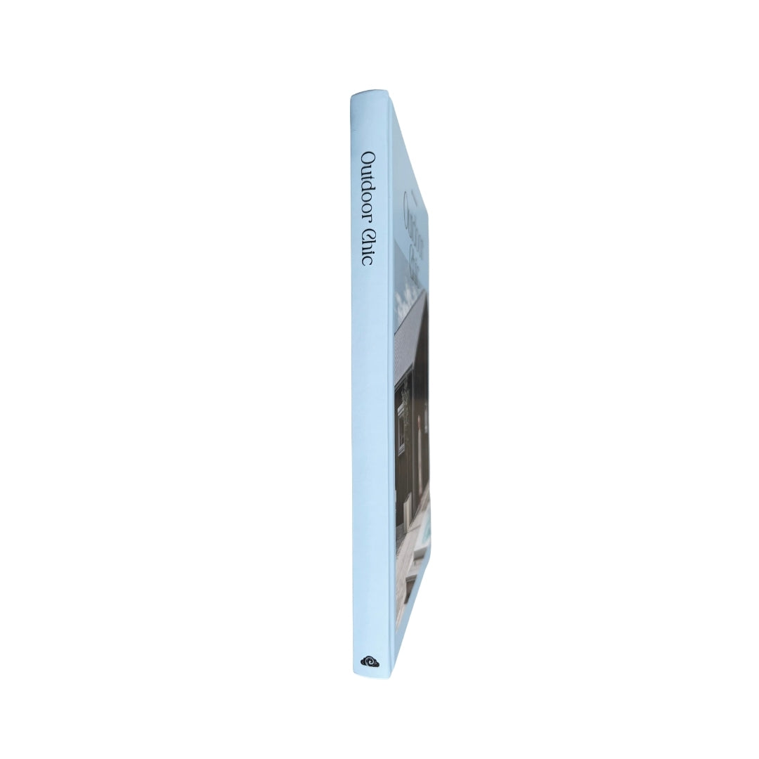 A single slim book or magazine standing upright on a plain white background, shown in side view. The spine is pale blue with the words 'Outdoor Chic' printed vertically, and the cover features a dark architectural photograph.