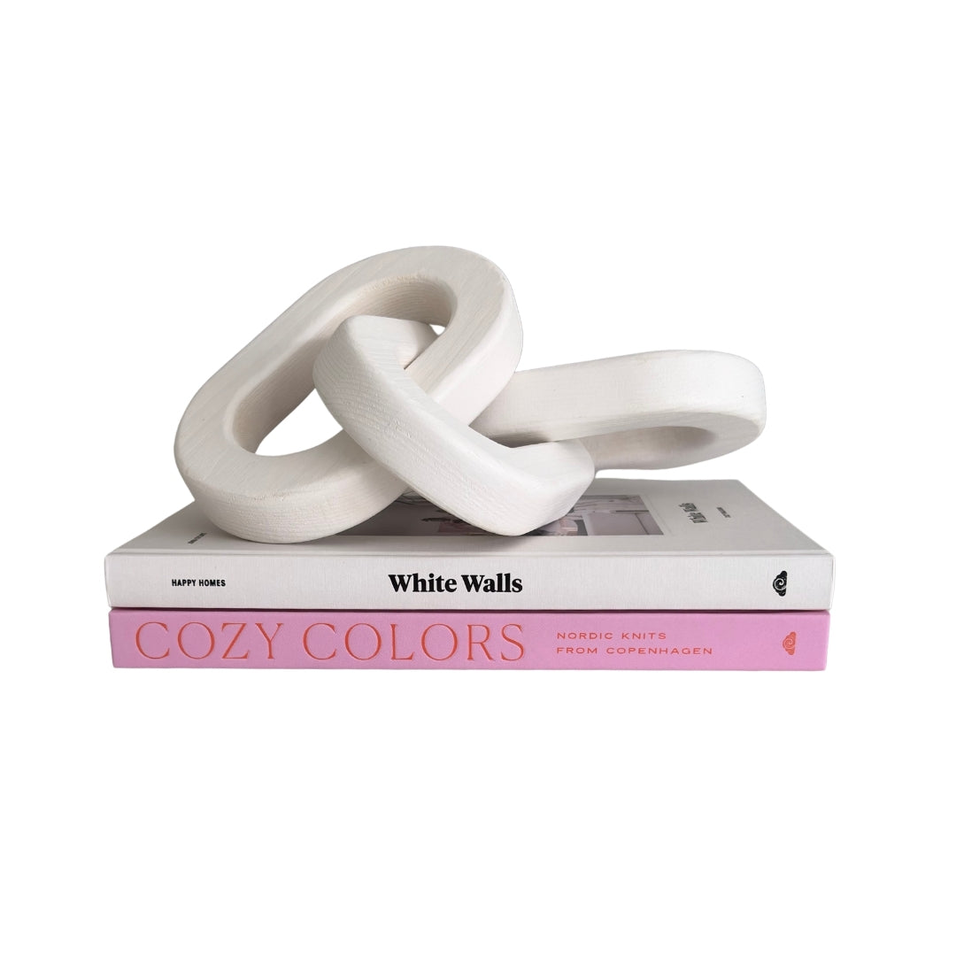 A white sculptural knot-like decorative object resting on top of two books, one white titled "White Walls" and one pink titled "COZY COLORS," against a plain white background.