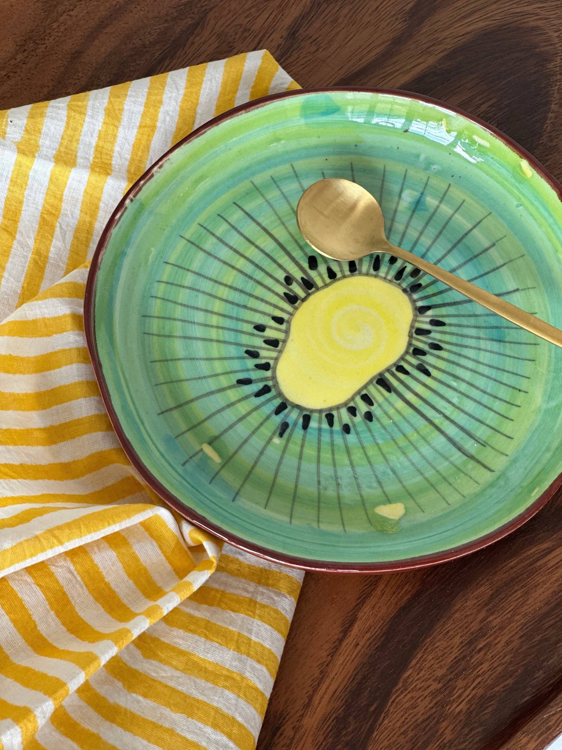Spring table setting featuring yellow striped napkins and Green ceramic plate with a kiwi slice and gold spoon on a wooden surface, modern table scape. 