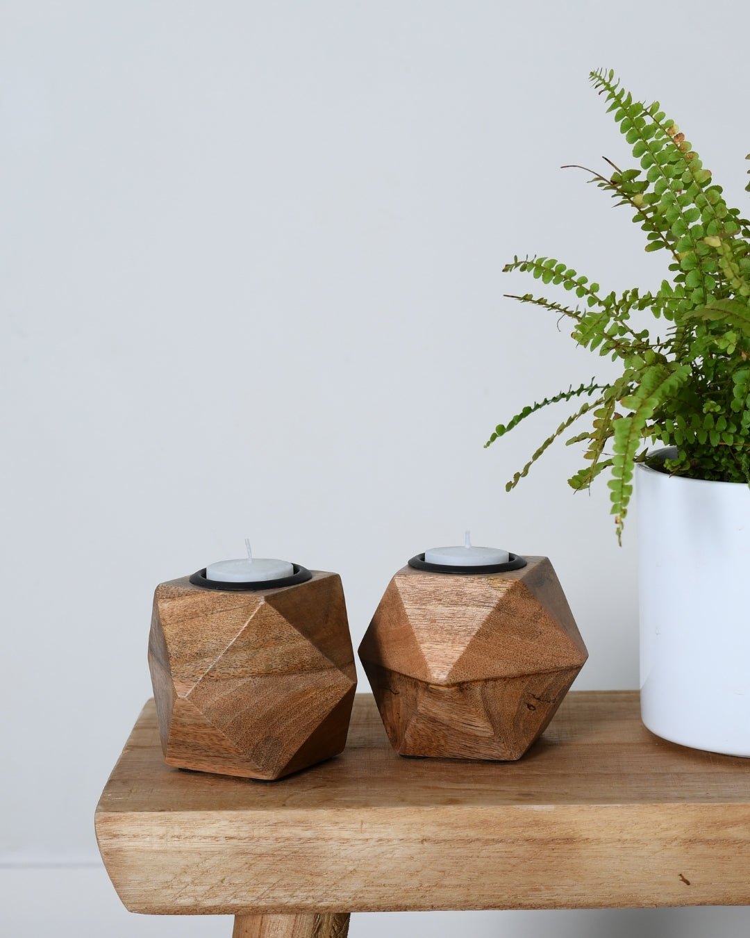 Two geometric wooden candle holders on a wooden table with a plant in the background.