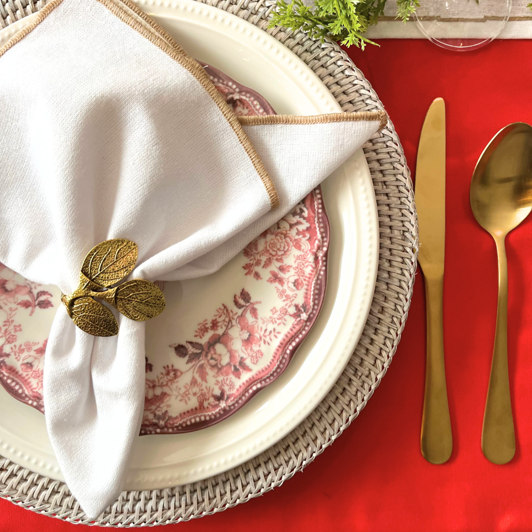 Dining table setting with floral plates, gold cutlery, and a white napkin with a leaf-shaped holder on a red tablecloth.