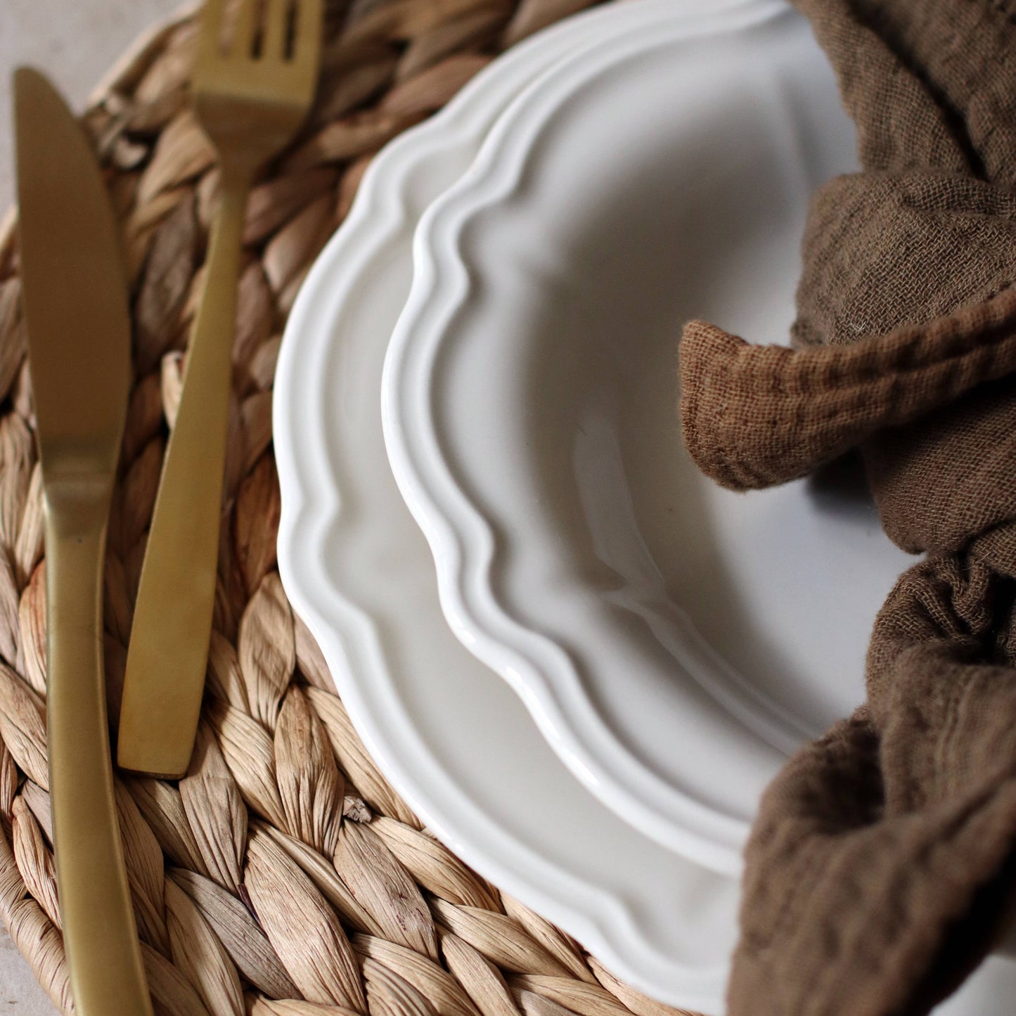 Close-up of White plate with gold fork and knife on a woven placemat, brown napkin beside