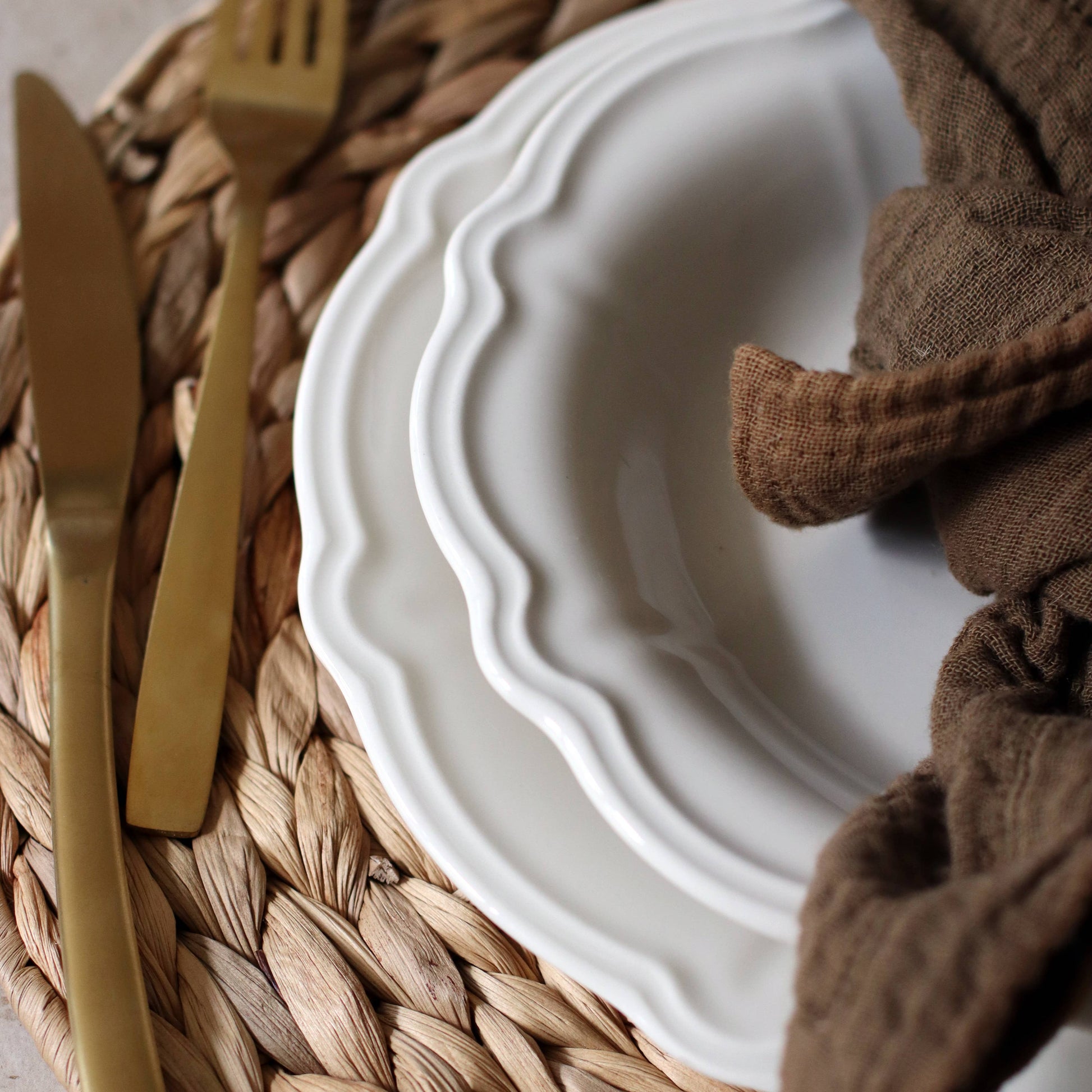 Close-up of White plate with gold fork and knife on a woven placemat, brown napkin beside