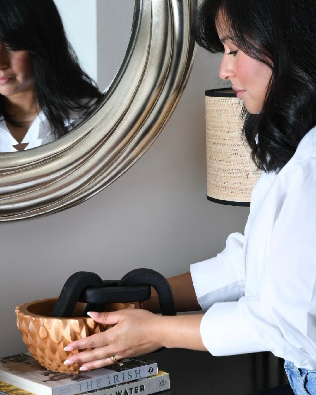 A woman in a white blouse places a black 3-Link Wood Chain Decor into a textured wood bowl, woven copper-colored bowl. The scene is set in a home environment with a large round silver-framed mirror on the wall, a woven lamp, and books on a table.