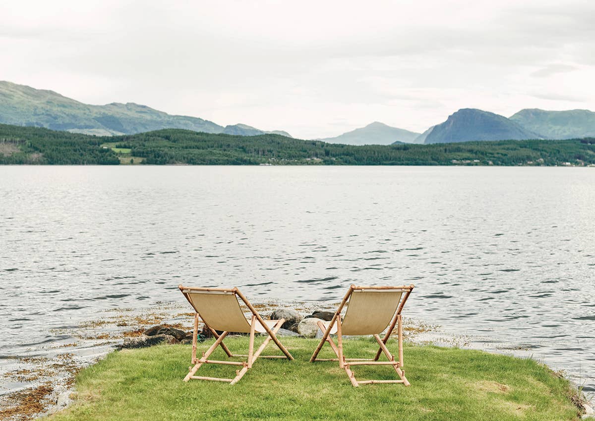 Two deck chairs on a grassy patch by a lake with mountains in the background