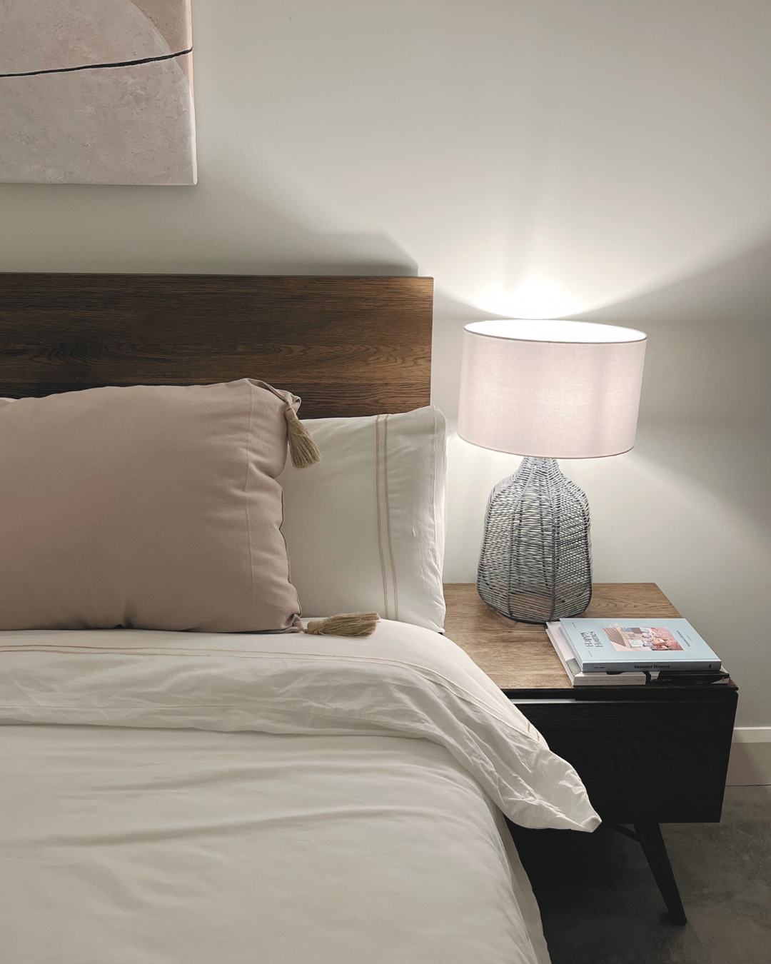 Bedroom with wooden headboard, white bedding, and a nightstand with a lamp and magazine. Sophisticated coastal bedroom sanctuary featuring a natural beige and soft stone palette bed pillow with rustic jute details.