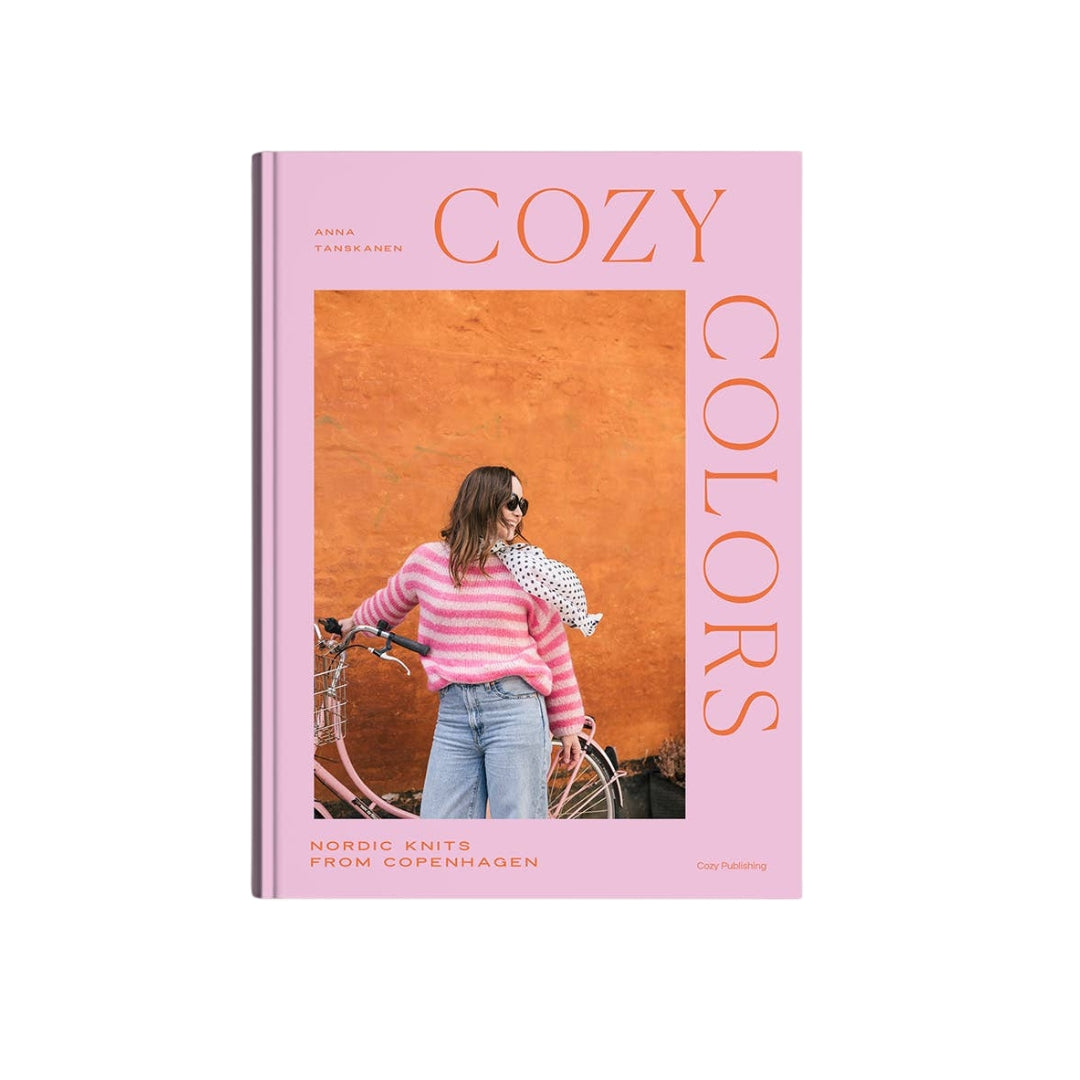 The book cover of 'Cozy Colors: Nordic Knits from Copenhagen' by Anna Fjerr Hansen, published by Care Publishing. The cover features a solid pink background with the title in large orange lettering and a central square photograph showing a woman in a pink striped sweater next to a bicycle against an orange wall.