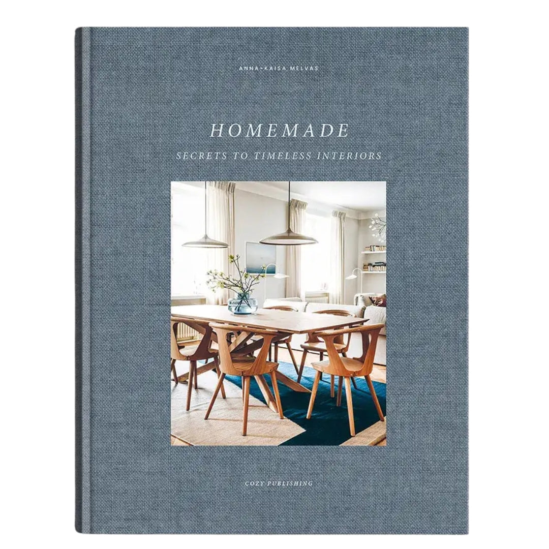 The front cover of the book 'Homemade: Secrets to Timeless Interiors' by Annabel Reese-Mellor, published by Good Facilities. The cover features a textured slate blue linen-like fabric background with a central color photograph of a mid-century modern dining room.