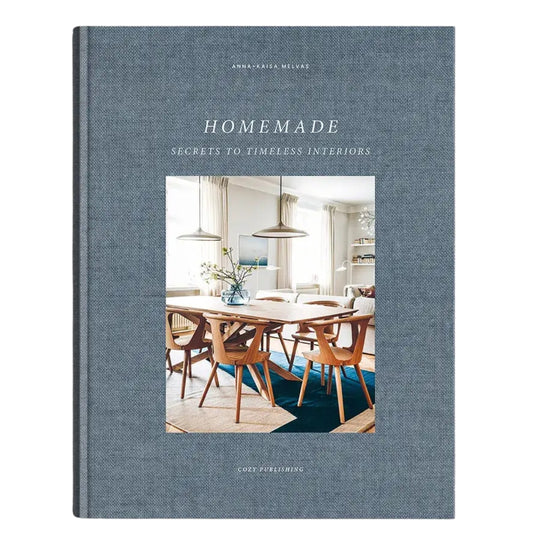 The front cover of the book 'Homemade: Secrets to Timeless Interiors' by Annabel Reese-Mellor, published by Good Facilities. The cover features a textured slate blue linen-like fabric background with a central color photograph of a mid-century modern dining room.