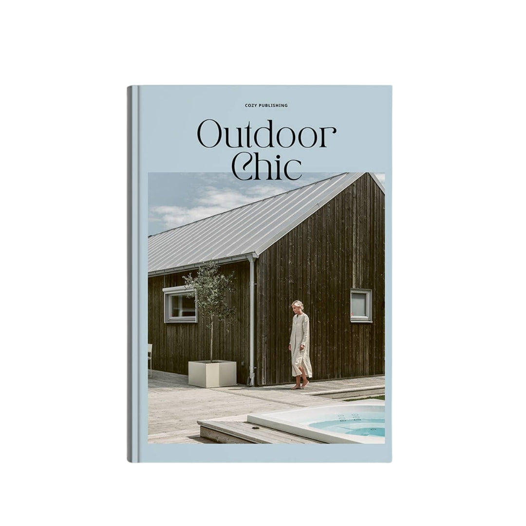 A hardcover book titled 'Outdoor Chic' by Fody Publications, featuring a light blue cover with a central photograph of a person in a long cream robe standing beside a dark wood cabin and a small pool.