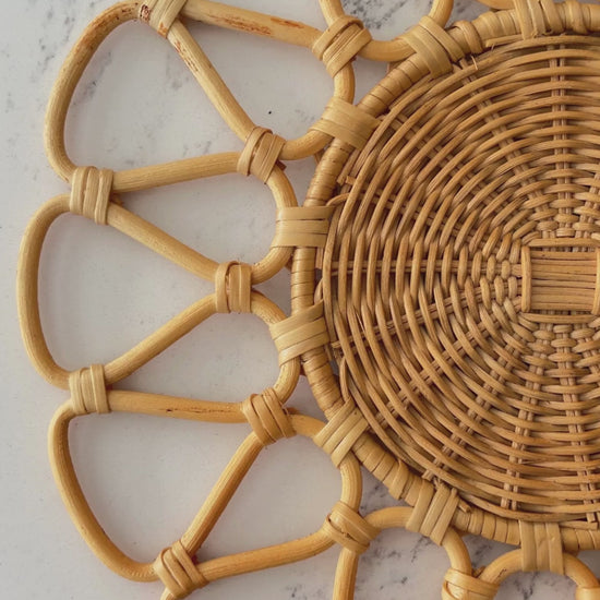 Cinematic slow-motion shot of a handwoven round natural beige rattan placemat with a sunburst floral pattern (D33cm).