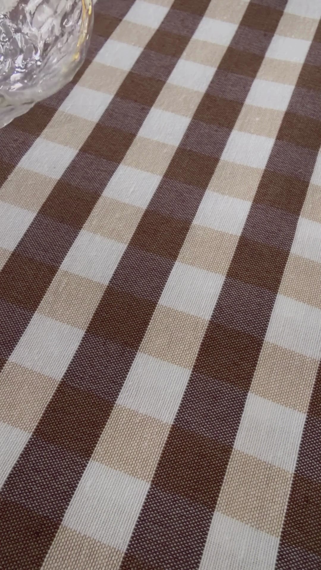 Close up demonstration of water being poured onto brown and beige gingham tablecloth fabric, forming droplets that can be wiped away.