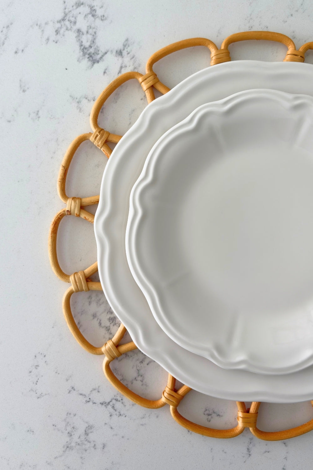 Two white ceramic dinner plates with scalloped edges stacked on top of a tan woven rattan placemat on light gray marble.